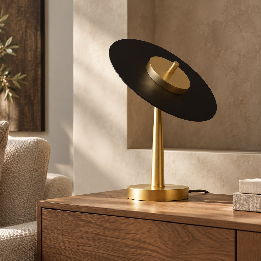 Lampe de table Anima à disque noir incliné et pied en laiton, sur un meuble en bois près d’un fauteuil.