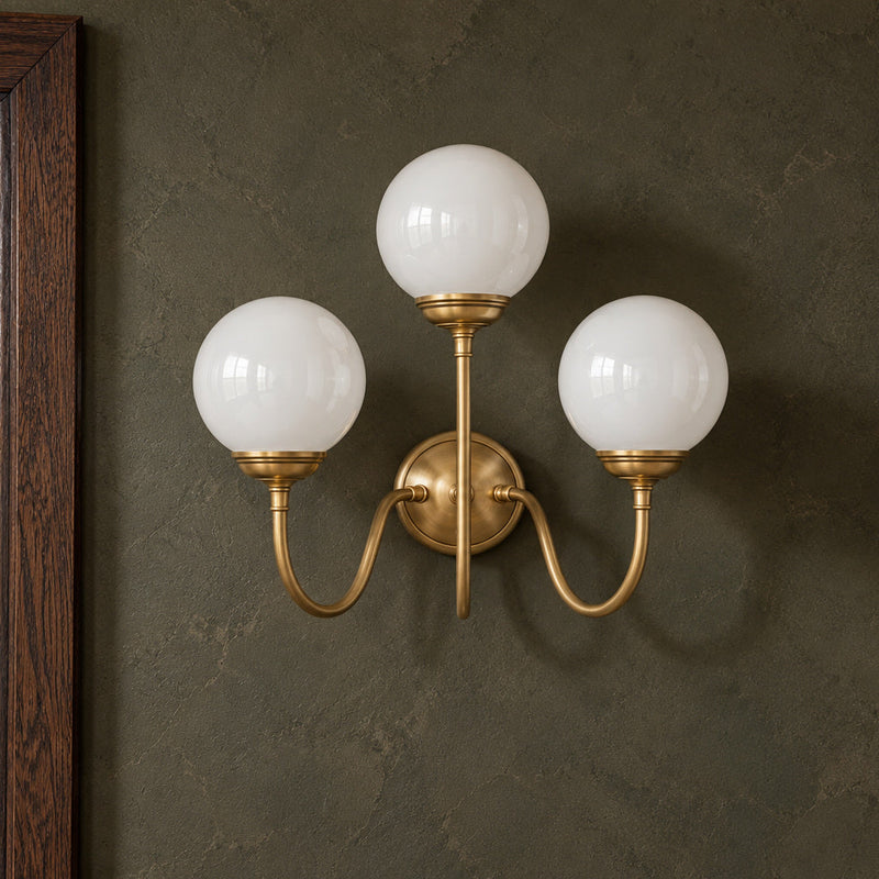 Incanto brass wall lamp with three glossy white globes beside a dark oak doorway.
