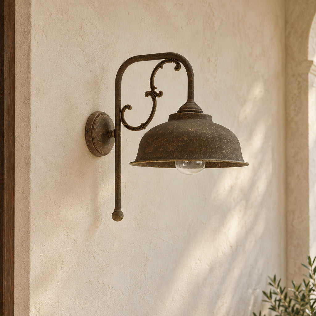 Applique Lipari en laiton vieilli, avec abat-jour en dôme et volutes décoratives, près d’une porte en bois sous une loggia.