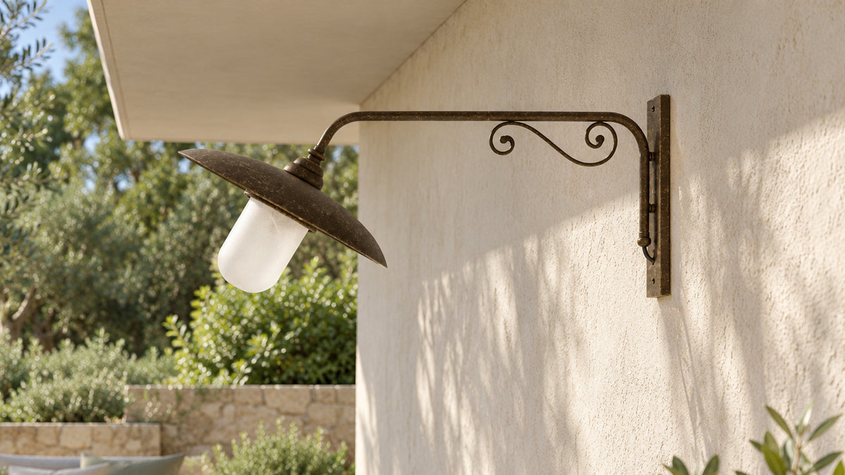 Cloe Wandleuchte aus Messing mit langem, von Voluten gestütztem Arm an einer sonnigen Loggiawand am Garten.